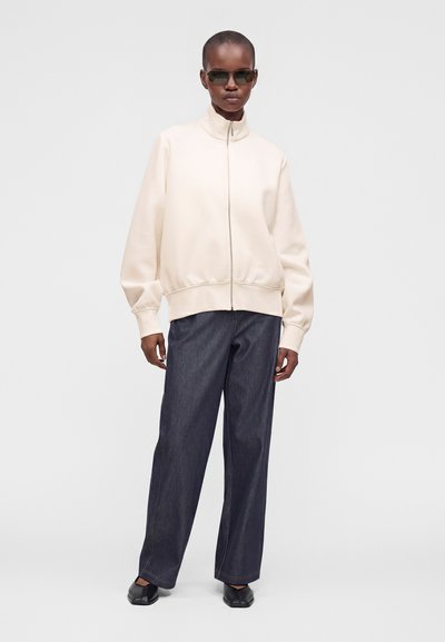 Personne portant des lunettes de soleil foncées, une veste crème zippée, un pantalon ample foncé et des chaussures noires à bout carré, debout devant un fond clair uni.
