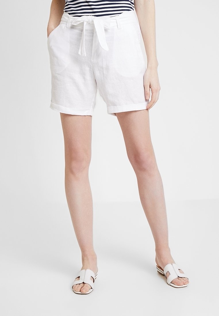 Pantaloni corti bianchi in lino con cintura annodata, dotati di tasche laterali e orlo arrotolato. Indossati con sandali in tela di colore chiaro.