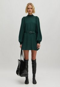 Grünes, langärmliges Kleid mit hohem Kragen und gerafften Schultern, tailliert mit einem passenden Gürtel. Schwarze kniehohe Stiefel und eine Tasche.