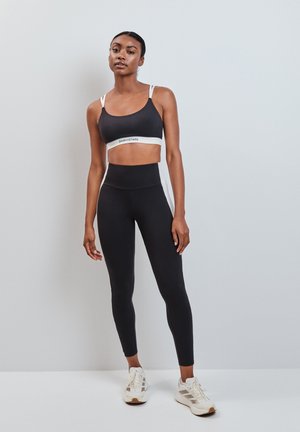Donna che indossa un reggiseno sportivo nero e leggings a vita alta con strisce bianche, in piedi davanti a uno sfondo grigio chiaro.