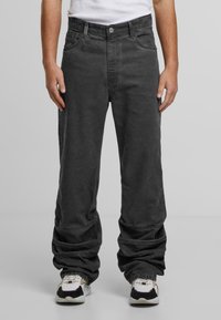 Muž v volných černých džínech a bílých teniskách, stojící s uvolněnýma rukama podél těla na jednobarevném šedém pozadí.