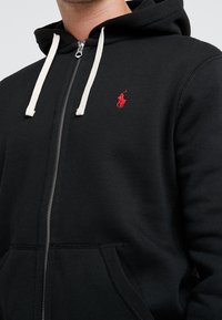Sudadera negra con cremallera fabricada en algodón. Cuenta con una gran capucha, cordones blancos y un pequeño logo rojo en el pecho. Textura suave.