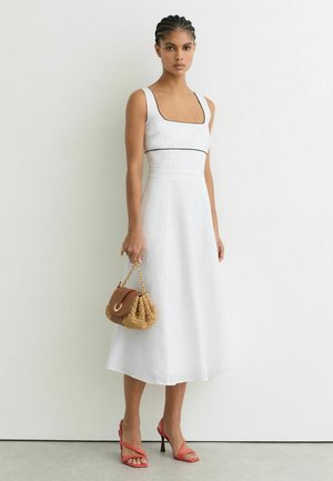 Frau trägt ein weißes ärmelloses Midikleid mit schwarzem Rand, hält eine braune gewebte Handtasche und trägt rote, geschlossene High-Heels mit Riemchen.