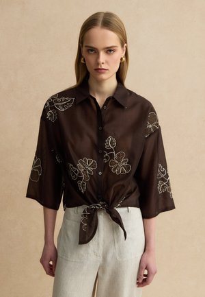 Jeune femme aux cheveux blonds raides portant une chemise marron foncé à motifs floraux nouée à la taille et un pantalon large crème, se tenant contre un mur beige.