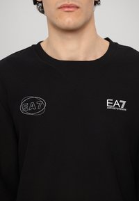 Črna dukserica z teksturirano tkanino, ki ima na prsih bele logotipe EA7 in Emporio Armani. Ima obliko V-izreza in ribana ovratnika.