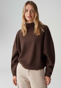 Bruine oversized trui met een hoge halslijn en doorgezakte schouders, gecombineerd met licht beige broek. Zachte textuur; casual, losse pasvorm.