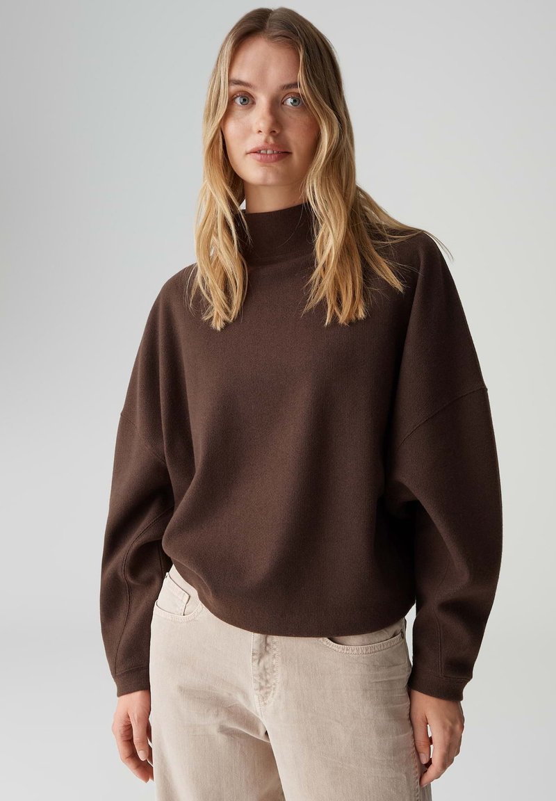 Bruine oversized trui met een hoge halslijn en doorgezakte schouders, gecombineerd met licht beige broek. Zachte textuur; casual, losse pasvorm.