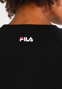 Camiseta de algodón negra con un cuello acanalado. Presenta un logo "FILA" en blanco y rojo en la parte superior de la espalda. Textura suave, diseño casual.