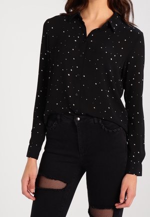 Woman wearing a black button-up shirt with white star patterns and black ripped jeans with fishnet fabric underneath.