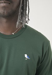 Grünes Baumwoll-T-Shirt mit einem kleinen, weiß-blauen gestickten Vogelmotiv auf der linken Brust. Weicher Stoff, runder Halsausschnitt.