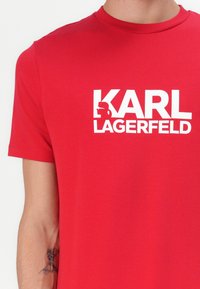 Raudona medvilninė marškinė su dideliu baltu grafiniu logotipu "KARL LAGERFELD" ir profilio ikona virš teksto. Trumpomis rankovėmis ir apvalia iškirpte.