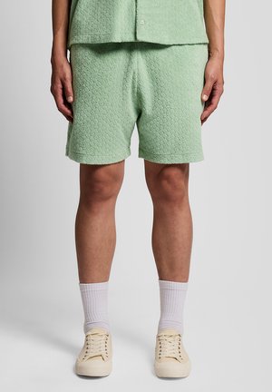 Mand iført teksturerede lysegrønne shorts og matchende skjorte, kombineret med hvide crew-sokker og beige snørebåndssko, stående på gråt gulv.