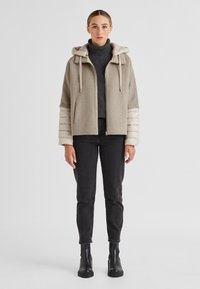 Beige Kapuzenjacke mit einem glatten, wolligen Oberteil, puffigen, cremefarbenen Ärmeln und einem Frontreißverschluss. Tragen mit dunkelblauen Jeans und schwarzen Stiefeln.