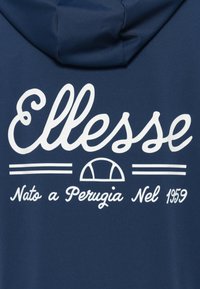 Hoodie bleu marine avec un logo en cursive blanc "Ellesse" et "Nato a Perugia Nel 1959" en dessous, présentant un design avec une ligne courbe et un demi-cercle.