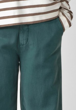 Inimese lähivõte, kes kannab türkiissiniseid pükse, käed taskutes, ja kreemikat ning pruunikat triibulist pika varrukaga särki.