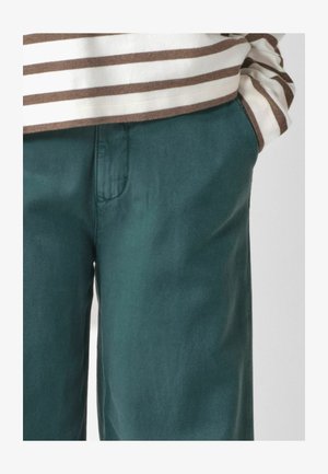 Inimese lähivõte, kes kannab türkiissiniseid pükse, käed taskutes, ja kreemikat ning pruunikat triibulist pika varrukaga särki.