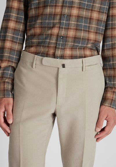Uomo che indossa pantaloni beige con chiusura nascosta e una camicia a maniche lunghe a quadri marrone, blu e beige. Mani rilassate ai lati.