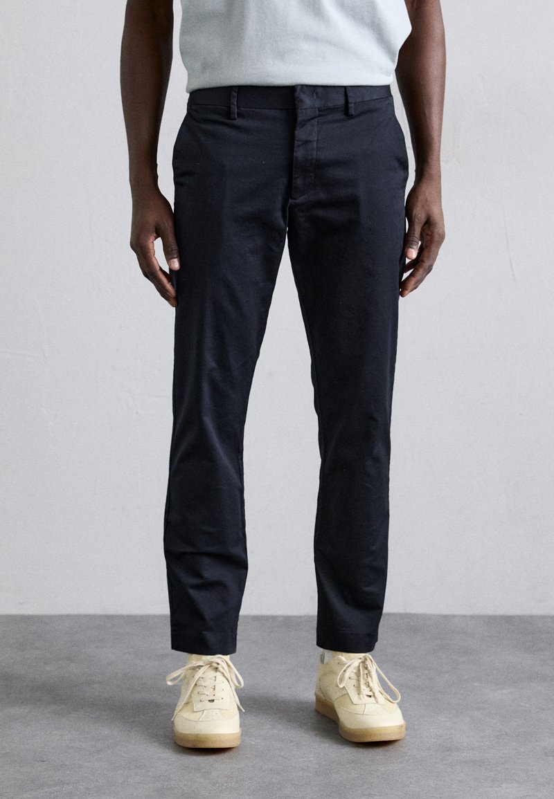 Pantalones azul marino de algodón, con un diseño de pierna recta, dos bolsillos frontales y construcción minimalista. Llevados con zapatillas claras.