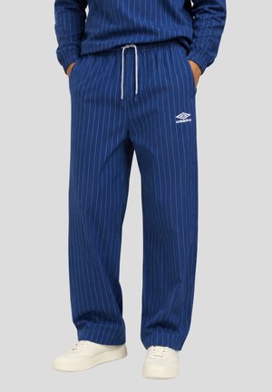 Pantalones azul marino con rayas finas, cordón blanco y logo de Umbro en el muslo izquierdo, usados con zapatillas blancas y manos en los bolsillos.