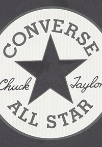 Kruhová grafika s texturovaným bílým pozadím a centrální černou hvězdou. Text zní "CONVERSE" a "Chuck Taylor All Star" v šedé barvě.