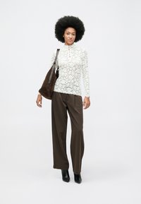 Top de encaje de manga larga en color crema claro, con cuello alto y botones decorativos, combinado con pantalones sueltos marrones y botas negras de tobillo.