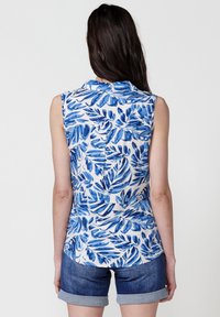 Blusa senza maniche con stampa di foglie blu e verde su sfondo bianco, collo e orlo arrotondato; abbinata a pantaloni shorts in denim.