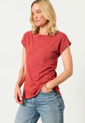 Blonde woman wearing a short-sleeve red T-shirt and blue jeans, with one hand in pocket and gold bracelets on wrist.