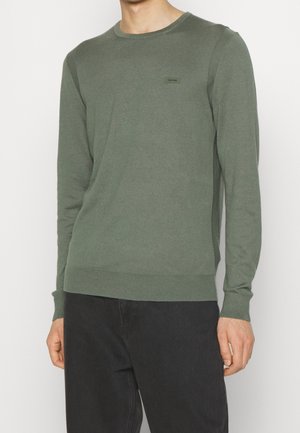 Homme portant un pull moulant vert olive à manches longues avec un petit logo Calvin Klein sur la poitrine et un pantalon noir.