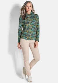 Langärmliges, grünes Oberteil mit Paisley-Muster und Stehkragen, kombiniert mit beigen Hosen und weißen Sportschuhen, vor einem neutralen Hintergrund stehend.