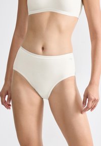 Vrouw in witte hooggeplaatste katoenen slips en bijpassende crop top, staande tegen een effen lichte achtergrond.