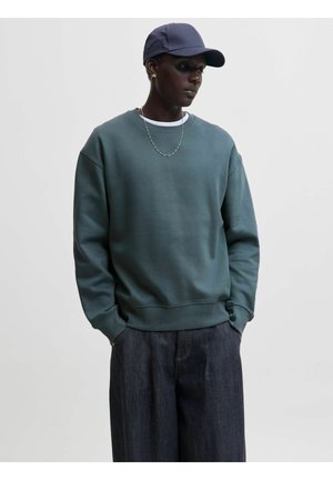 Osoba nosząca ciemnozielony sweatshirt, ciemne szerokie dżinsy, granatową czapkę i srebrny łańcuszek na szyi, patrząca w dół z rękami w kieszeniach.