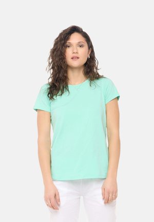 Jonge vrouw met krullend bruin haar, die een muntgroen T-shirt met korte mouwen en een witte broek draagt, staand tegen een effen lichte achtergrond.