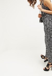 Mujer con un mono estampado en blanco y negro, sandalias de tacón negras con borlas, sosteniendo un pequeño bolso negro con cadena.