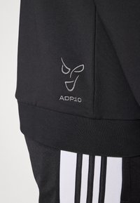 Μαύρο αθλητικό φούτερ με ασημένιο κεντημένο λογότυπο και κείμενο "ADP-10", με ριμπ μανσέτες και κομψή υφή. Περιλαμβάνει λευκές ριγέ λεπτομέρειες.