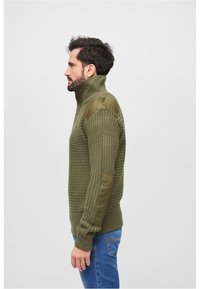 Olivno zelen pulli iz pletenine z visokim ovratnikom, rebrasto teksturo in podloženimi rameni. Ima tri gumbe in oprijete manšete.