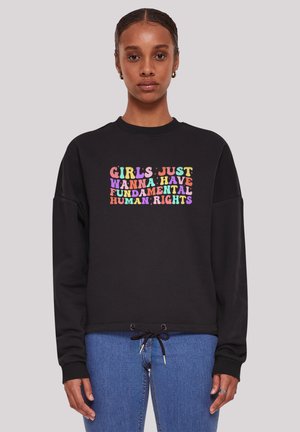 Frau trägt einen schwarzen Pullover mit buntem Text: "Mädchen wollen einfach nur grundlegende Menschenrechte haben", kombiniert mit blauer Jeans.