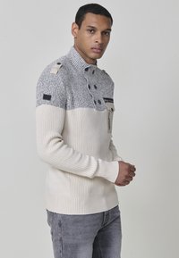 Suéter de punto en crema y gris, con un alto cuello, detalles de botones y un diseño de canalé texturizado. Incluye un detalle de bolsillo en el pecho.