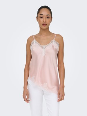 Donna che indossa un camisole in raso rosa pallido con bordo in pizzo bianco, abbinato a pantaloni bianchi, in piedi davanti a uno sfondo chiaro e uniforme.