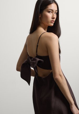 Junge Frau mit langen dunklen Haaren, die ein ärmelloses dunkles Kleid mit einer großen Schleife am offenen Rücken trägt.