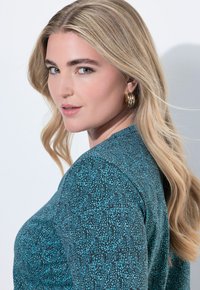 Turkosmönstrad topp med mjuk textur och rund halsringning. Modellen visar långt, vågigt hår och stora guldörhängen.