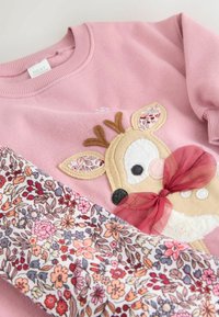 Sweatshirt roz cu aplicație de ren și accente cu model floral. Include detalii texturate și o fundiță dintr-un material moale și transparent.