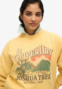 Felpa gialla con un design grafico che presenta "SuperDry" e "Joshua Tree National Park." Ha un colletto a costine e una texture morbida.