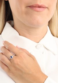 Donna con camicia bianca che mostra un anello d'argento con una pietra ovale blu scuro sulla mano sinistra appoggiata sul petto.