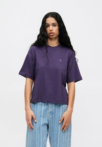 Violeta kokvilnas krekls ar apaļu izgriezumu, īsām piedurknēm un smalku logo uz krūškurvja. Saskaņots ar gaiši zilām svītrainām biksēm.