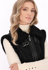 Zwarte nepbont vest met een ritsluiting en gespdetails, gedragen over een crèmekleurige geribde coltrui. Gladde en gemengde materialen.