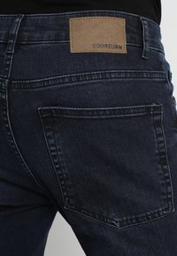 Jeans de mezclilla azul oscuro con un bolsillo trasero y una etiqueta de cuero marrón con la inscripción "YOURTURN" en la cintura.