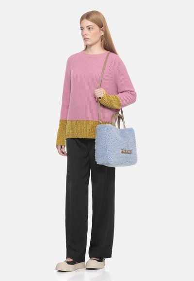 Maglione lavorato a maglia di colore rosa con dettagli dorati, abbinato a pantaloni neri e una borsa a tracolla soffice di colore azzurro chiaro con logo. Scarpe con dettaglio nero.