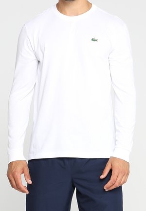 Man draagt een eenvoudige wit lange mouwen shirt met een klein groen krokodillenlogo op de borst en donkerblauwe broek, staand met ontspannen handen.