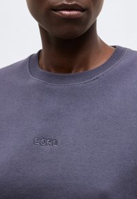 Κοντινό πλάνο ενός ατόμου που φοράει μωβ φούτερ με στρογγυλή λαιμόκοψη, με την επιγραφή "BORG" κεντημένη στο στήθος.