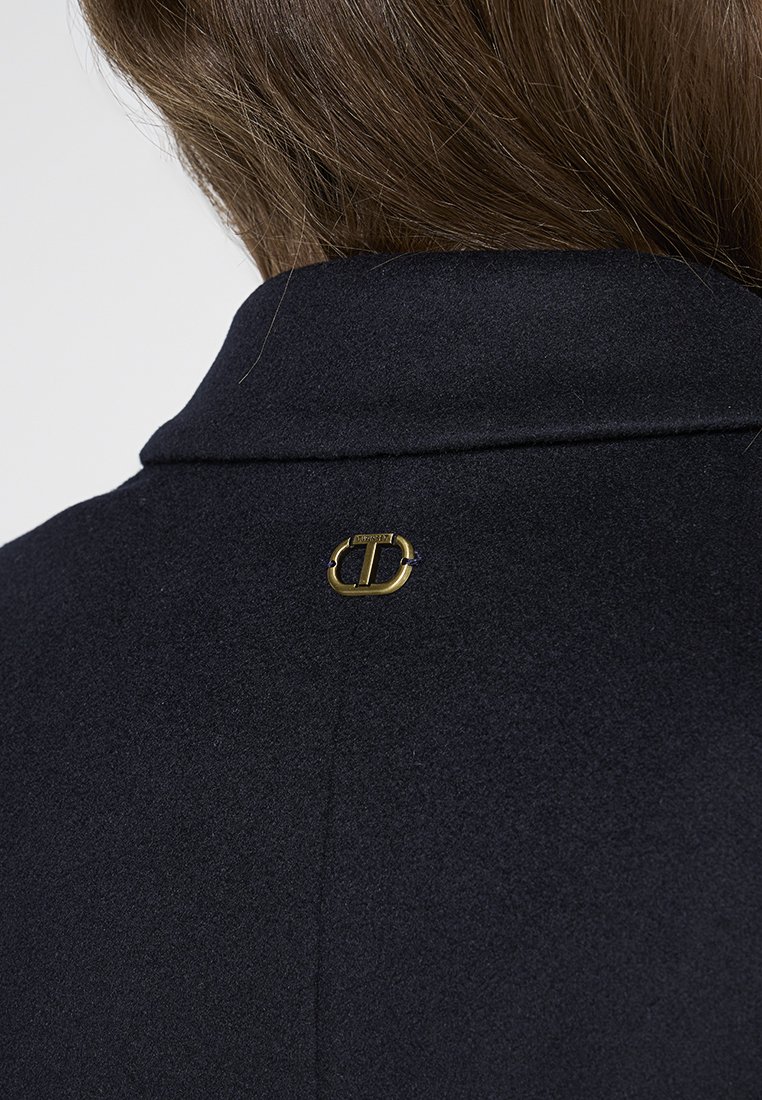 Bruin haar boven de kraag van een marineblauwe wollen jas met een klein gouden merkembleem in het midden van de bovenrug.